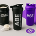ABE shaker standing upright on a countertop, next to an open tub of protein (either Applied Nutrition or MSS-branded), with a scoop of powder ready to go. To one side, a bold MSS-purple shaker brings in that unmistakable pop of colour.