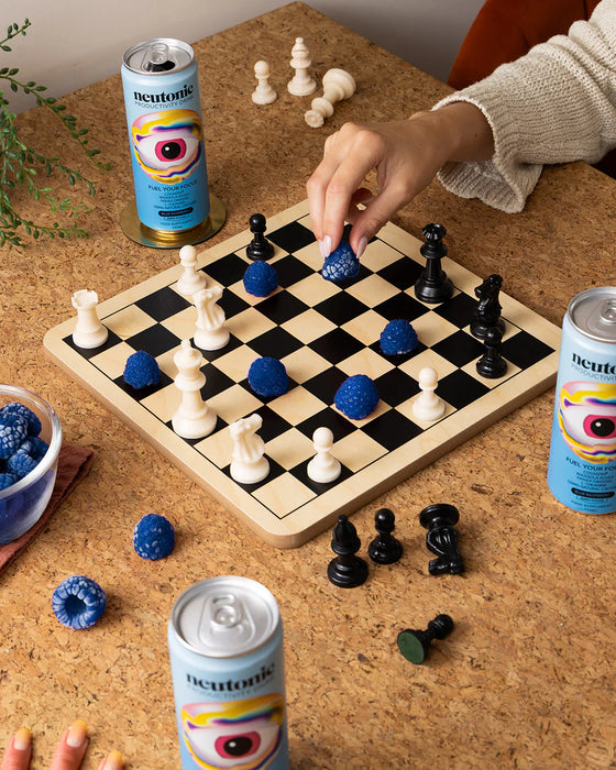 Person playing chess with blue and black pieces on a checkered board, surrounded by Heineken cans and a bowl of berries.