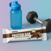 PER4M Protein Bar with blue water bottle and dumbbells on gradient MSS color background – Post-Workout Bite lifestyle image for MySupplementShop ecommerce listing.